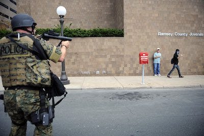 A police officer is pointing a green rubber-bullet gun at a protester who has his hands up and is on one knee