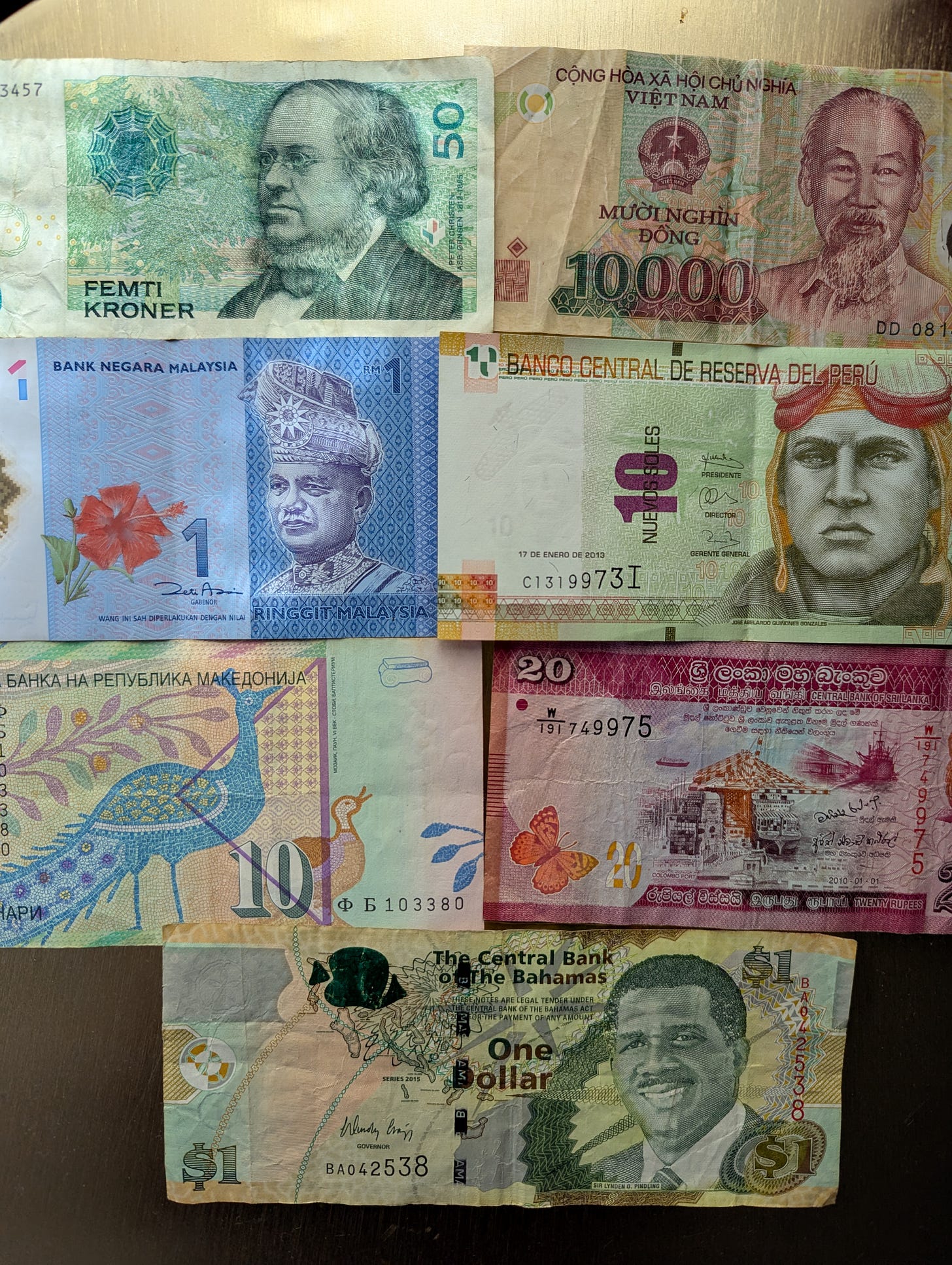 A collage of various international paper currencies laid out in a grid format. The currencies are from different countries and vary in color, design, and denomination. The denominations visible include 10,000 Vietnamese Dong, 50 Swedish Kroner, 1 Malaysian Ringgit, 10 Peruvian Nuevos Soles, 10 Macedonian Denars, 20 Sri Lankan Rupees, and 1 Bahamian Dollar. Each currency features a portrait or national symbol, along with text in the respective language.
