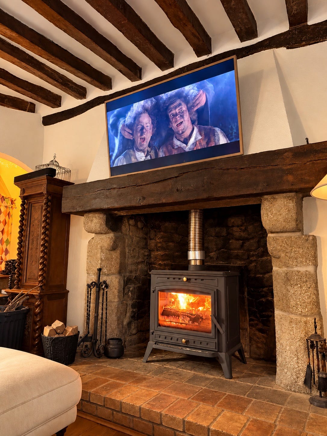 A cast-iron wood stove burning brightly inside a huge old stone fireplace with carved stone sides and a massive dark wooden beam as a mantel. A television above shows two cheering characters from a fantasy film. A carved wooden cabinet with barley-twist columns and a basket of firewood stand to the left. A cast-iron wood stove burning brightly inside a huge old stone fireplace with carved stone sides and a massive dark wooden beam as a mantel. A television above shows two cheering characters from a fantasy film. A carved wooden cabinet with barley-twist columns and a basket of firewood stand to the left.