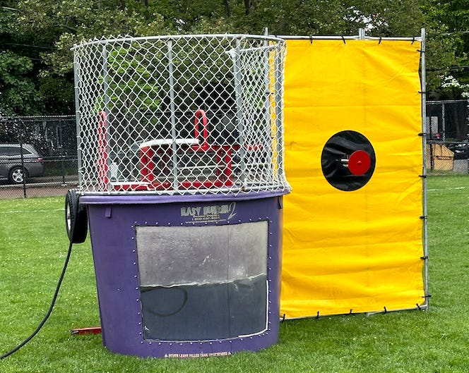 A portable dunk tank, half-full. to the right, is the target area, backed by a yellow tarp.