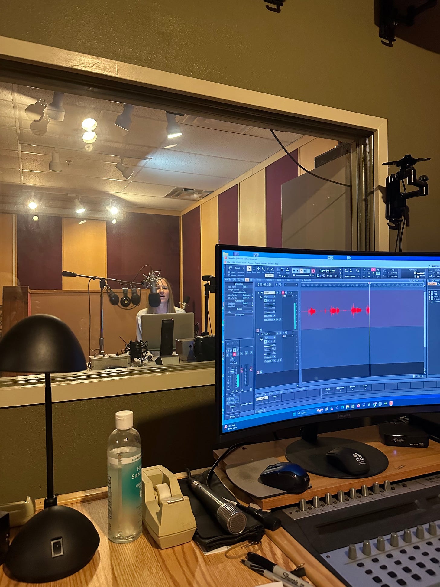 Photo of me sitting inside the sound recording booth. I'm behind a glass window speaking into a microphone, and the sound engineer's computer is in the foreground with sound waves across a recording screen.