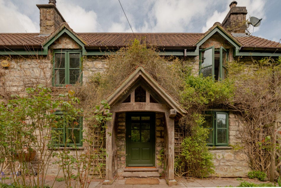 A light stone cottage with green timber trim and window frames and lots of foliage. It looks like somewhere Miss Honey could live