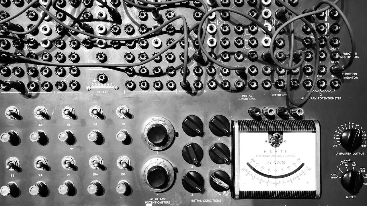 GPT-4o (edited): A black-and-white photo of a vintage analog computing system with numerous wires, dials, switches, and input jacks. The upper portion is dominated by tangled cables plugged into various ports, while the lower section features knobs and gauges, including a voltmeter labeled “HEATH ELECTRONIC ANALOG COMPUTER” and “D.C. Volts.” The setup suggests an intricate, manual process of calibration and configuration, evoking a sense of early technology and hands-on interaction.