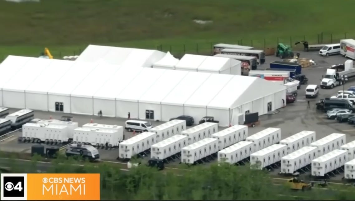 aerial screenshot of part of the tent city prison camp, with a large white tent, multiple prefab cells on trailers, and assorted law enforcement vehicles parked around the structures 