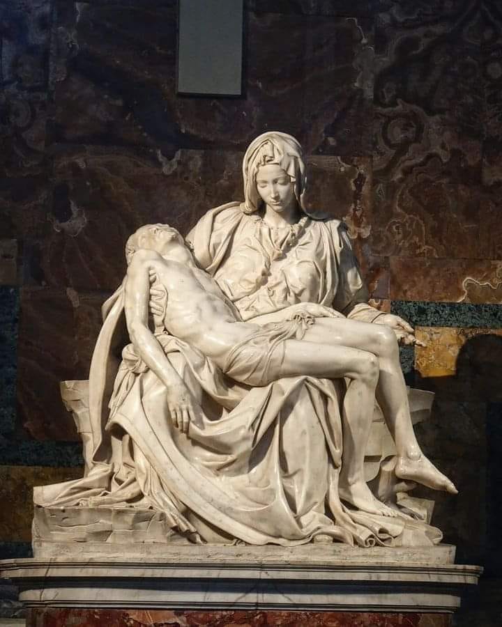 The image shows Michelangelo's famous sculpture, the Pietà, which depicts the Virgin Mary holding the body of Jesus after his crucifixion. The sculpture is intricately detailed, with Mary's expression conveying deep sorrow and tenderness. The post text reveals that Michelangelo was only 23 years old when he created this masterpiece, adding to the awe of his youthful genius. The sculpture is set against a backdrop of richly colored marble, enhancing its solemn and sacred atmosphere.