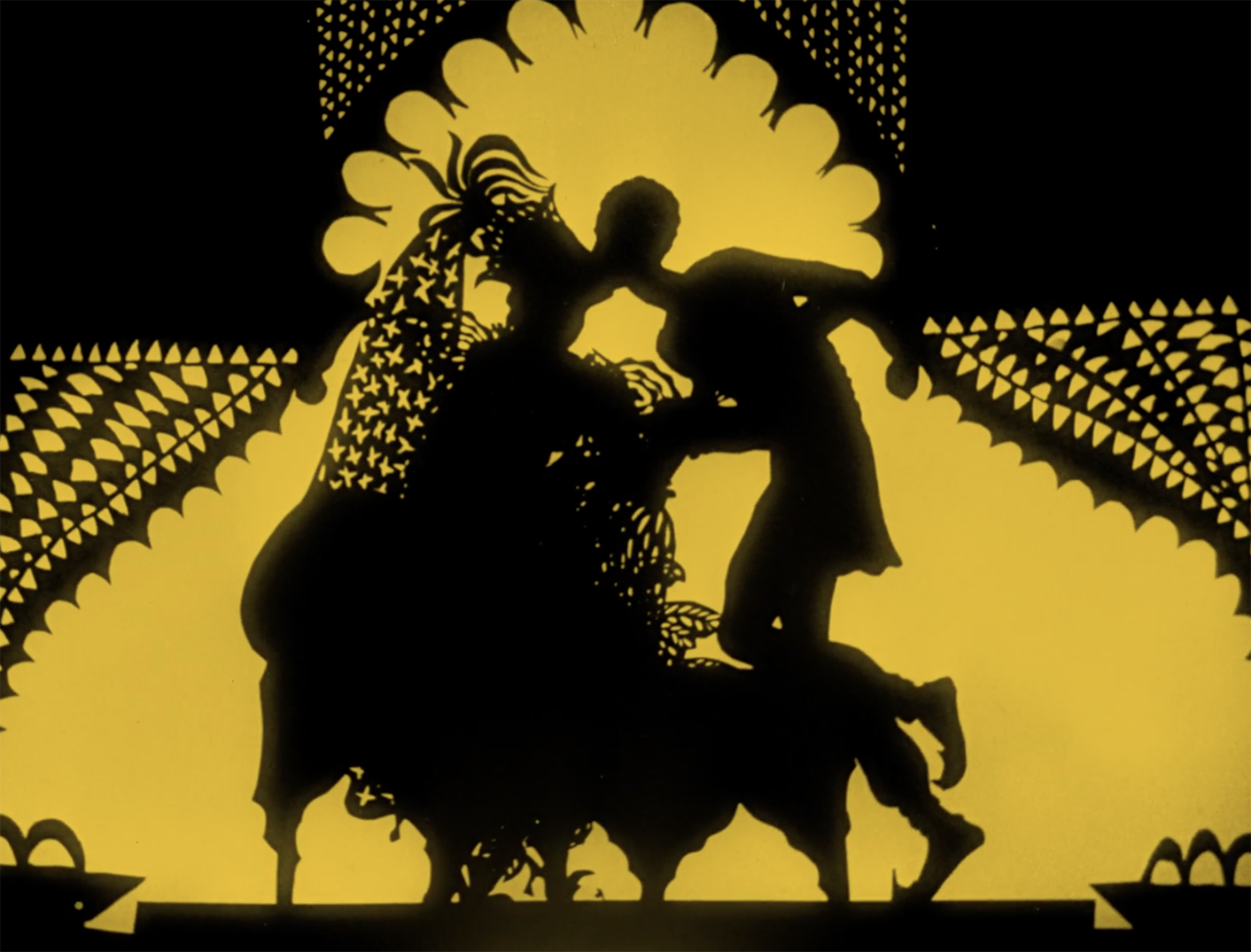 A still image from Lotte Reiniger's 1926 animated feature, The Adventures of Prince Achmed. Two silhouetted and lavishly dressed figures are kissing against a solid golden background.