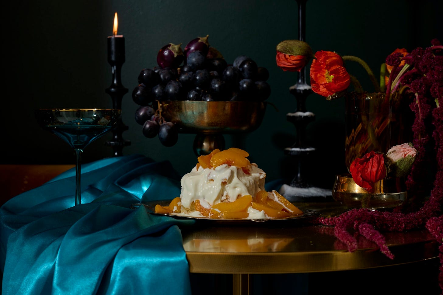 Peaches & Cream Cake topped with syrupy peaches and draped in whipped amaretto cream, styled on a gold table with teal satin, black grapes in a brass bowl, candlelight, and vibrant red poppies in the background.