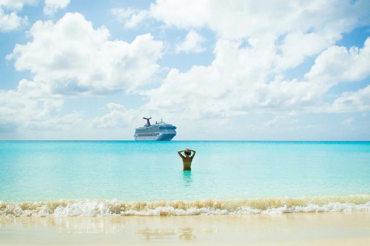 A person stands waist-deep in turquoise water facing a large cruise ship on the horizon beneath a bright blue sky filled with clouds. Gentle waves lap at the sandy shore in the foreground.