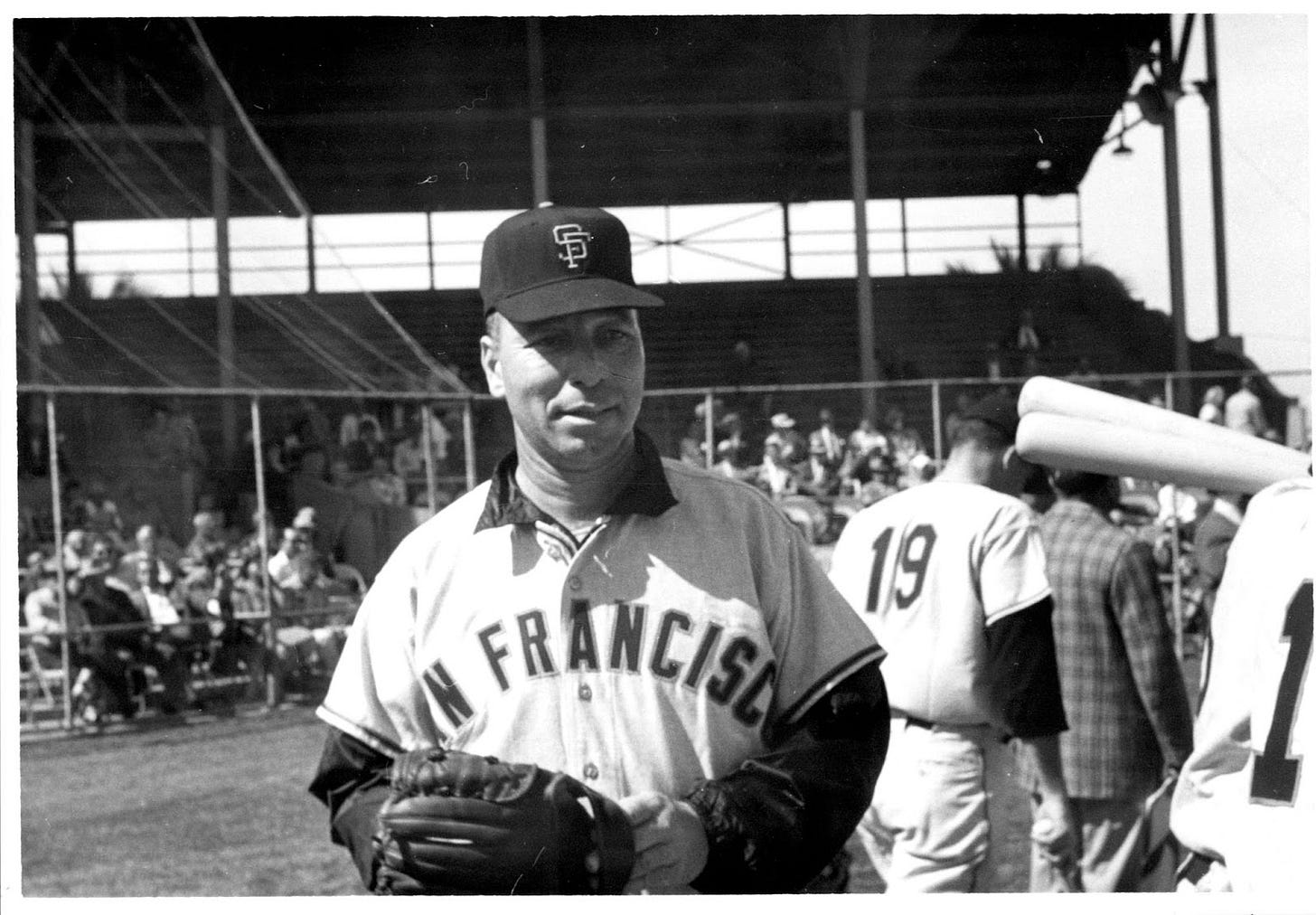 Sam “Toothpick” Jones is shown in a black and white photo wearing a white SF Giants jersey, with a long sleeve coaches jacket underneath and baseball glove. He stands on the field with fans and players behind him. He has a focused facial expression where his light complexion is hidden under the shadow on his black SF Giants baseball cap.