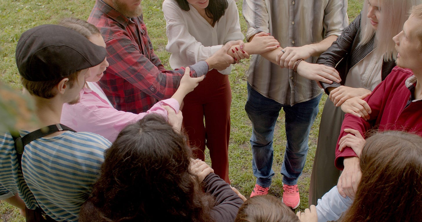 With no faces visible, this is a photo of 10 people outdoors in a circle holding hands and forearms. Clearly a support group of some sort. Communal healing.