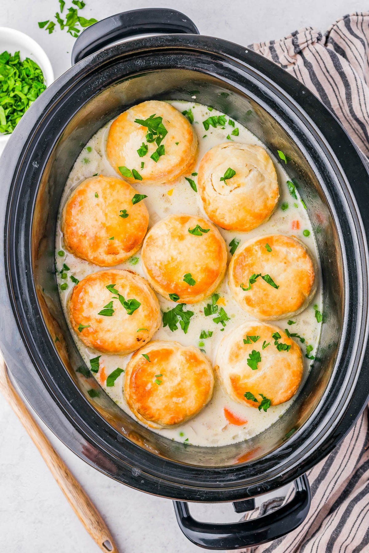 Slow cooker with creamy chicken stew topped with golden biscuits and garnished with chopped parsley.