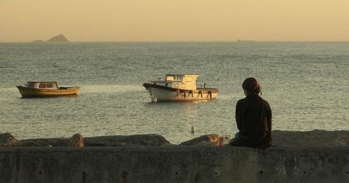 This may contain: a man sitting on the edge of a wall looking out at boats in the water
