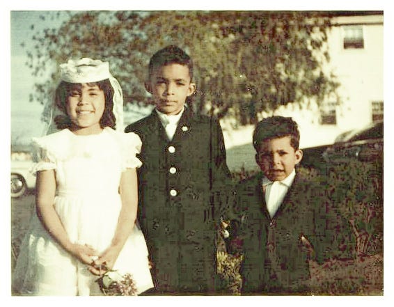 A color photo of three children standing outdoors. A girl wearing a white First Communion dress with a veil and gloves stands on the right. Two boys stand beside her in dark suits with white shirts and ties.