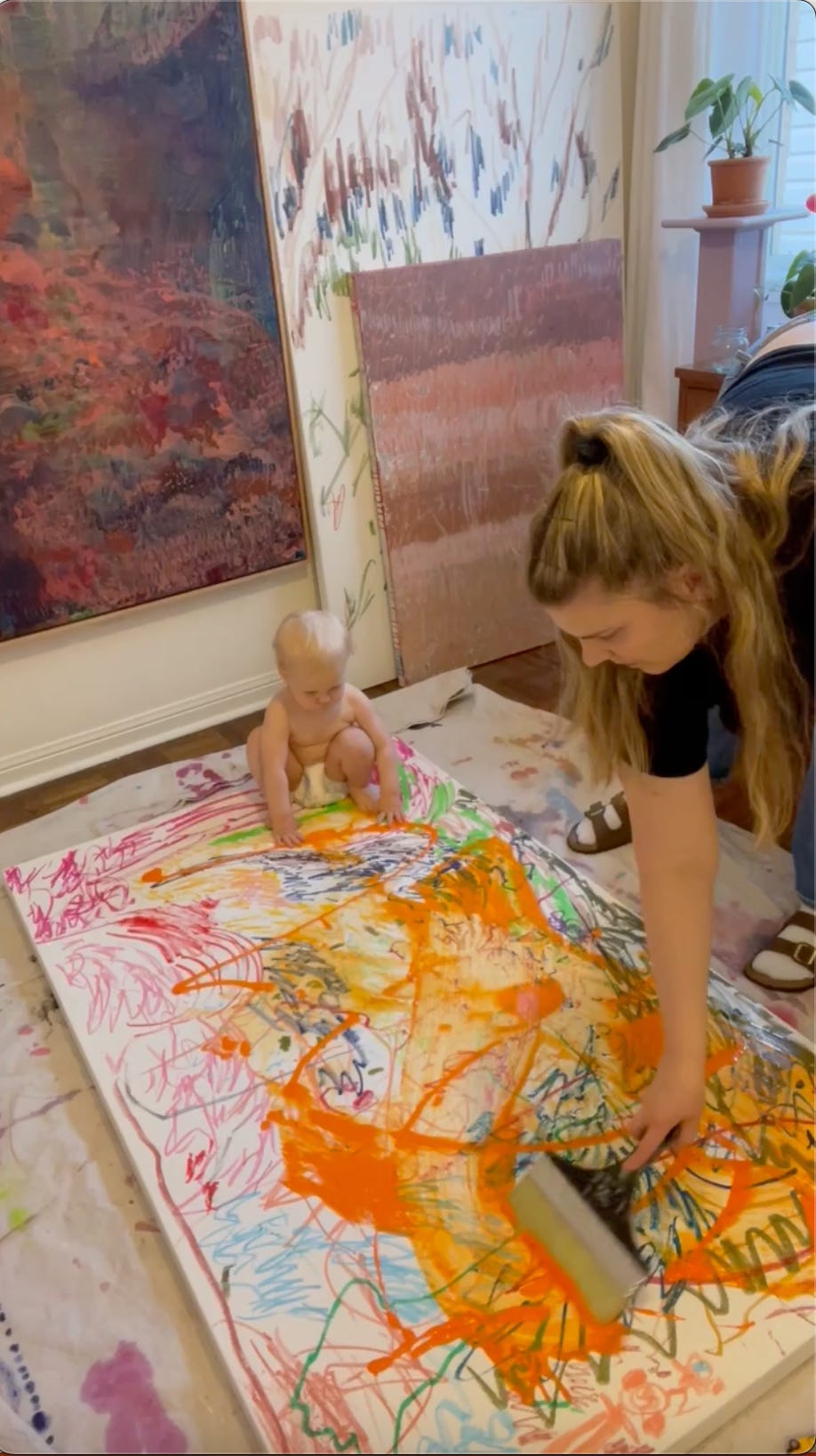 Adult pushing watery orange paint with a brush while infant sits on the canvas (underpainting for Liquid Light).