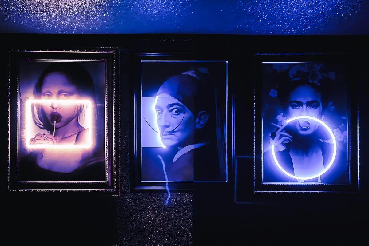 Three framed art prints, including portraits of Salvador Dali and Frida Kahlo, illuminated by custom purple and white neon light tubes in a dark room. Three framed art prints, including portraits of Salvador Dali and Frida Kahlo, illuminated by custom purple and white neon light tubes in a dark room.