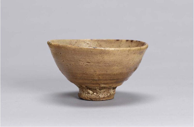 [AI Generated Alt Text] Small footed ceramic tea bowl with beige-brown glaze and rough textured base on a light gray background