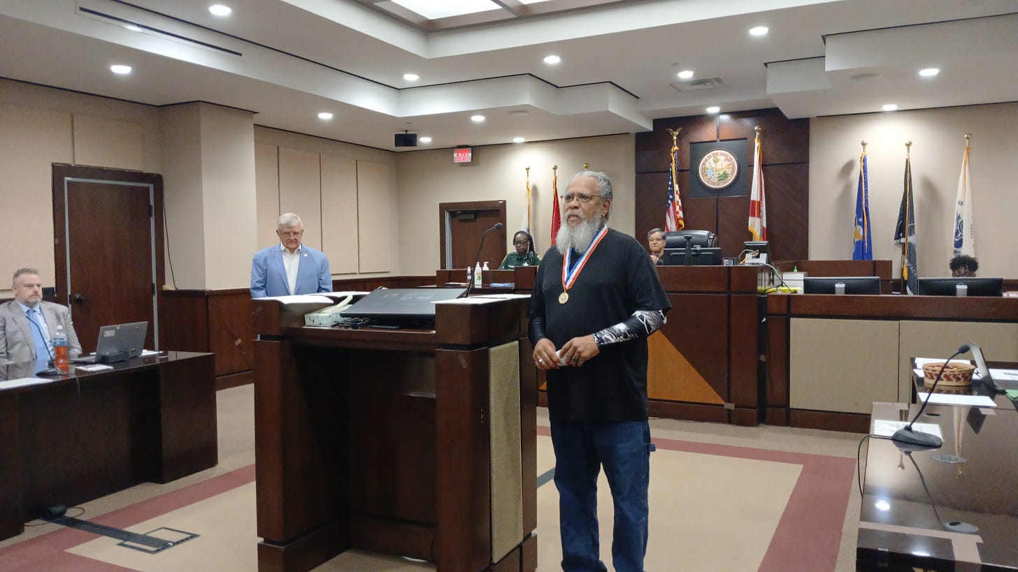 The image shows the interior of a courtroom with several individuals present. A man with a white beard, wearing a black shirt and a medal around his neck, stands in the foreground near a podium. Behind him, people are seated at desks and the judge's bench, including a man in a light blue suit and others in business attire. The background displays various flags, including the American flag and state flags, and a seal on the wall. The setting appears formal, likely during a ceremony or official proceeding. No specific names, recognizable figures, or brands are visible.