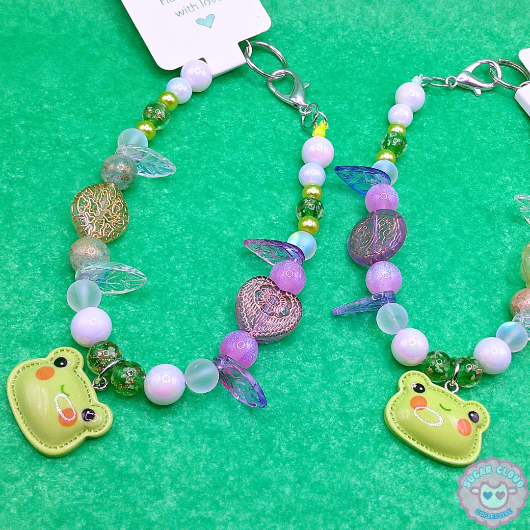 Two different styles of "The Meadow's Muse" wristlet keychain handmade with a variety of white, glitter, gold, and green beads handmade by Strawberry Erin. Both styles feature an adorable acrylic charm of a frog face outlined with a hand-stitch pattern.