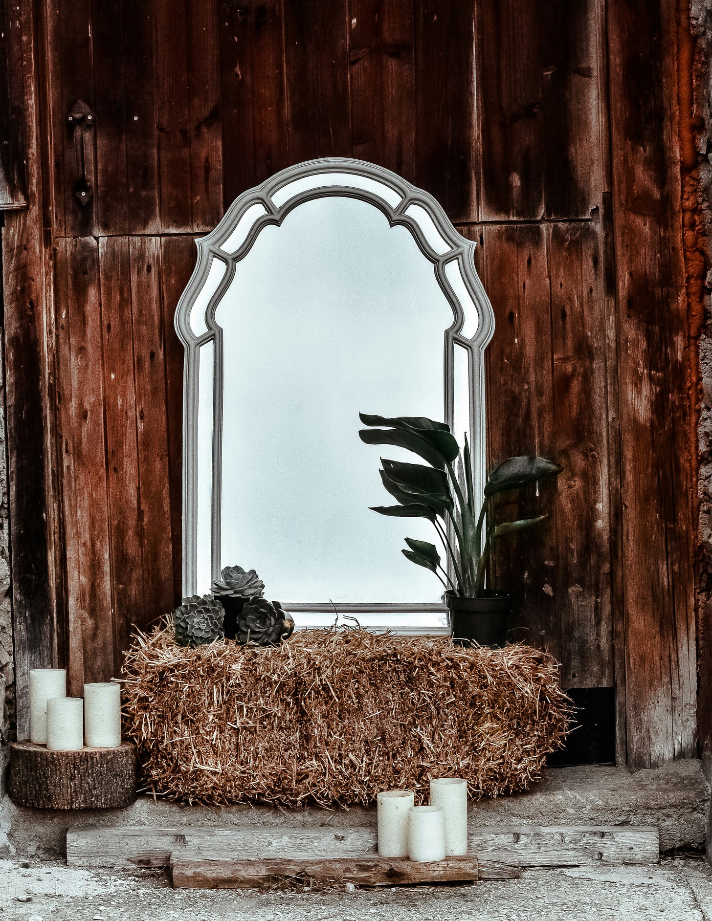 Adorned mirror sitting on a pile of hay, leaving against a wooden barn wall, with dry plants and candles around.