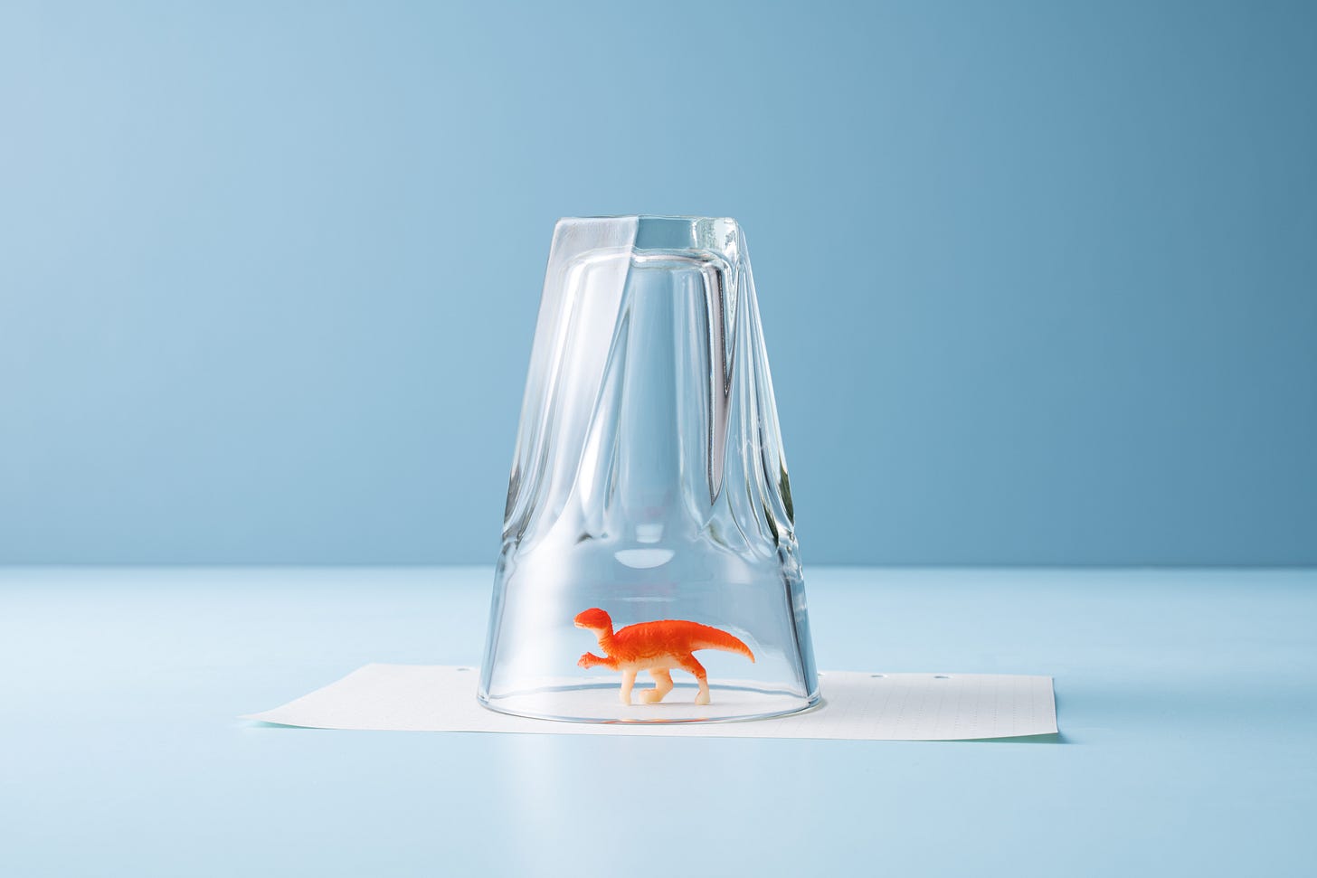 Orange toy dinosaur under an upside down drinking glass on top of a piece of paper, almost like it was a real creature captured to  be set free outside