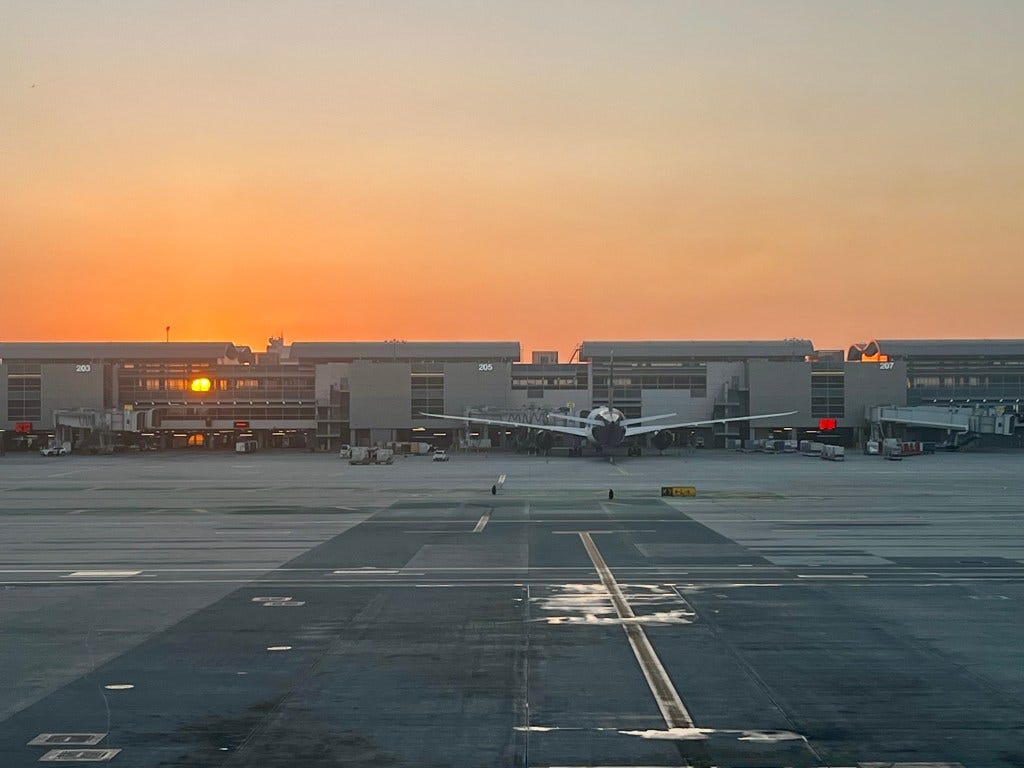 At Los Angeles Airport, the glowing sun burns through concourse windows across an apron of pavement, a commercial airplane docked. The hazy sky is lit orange.