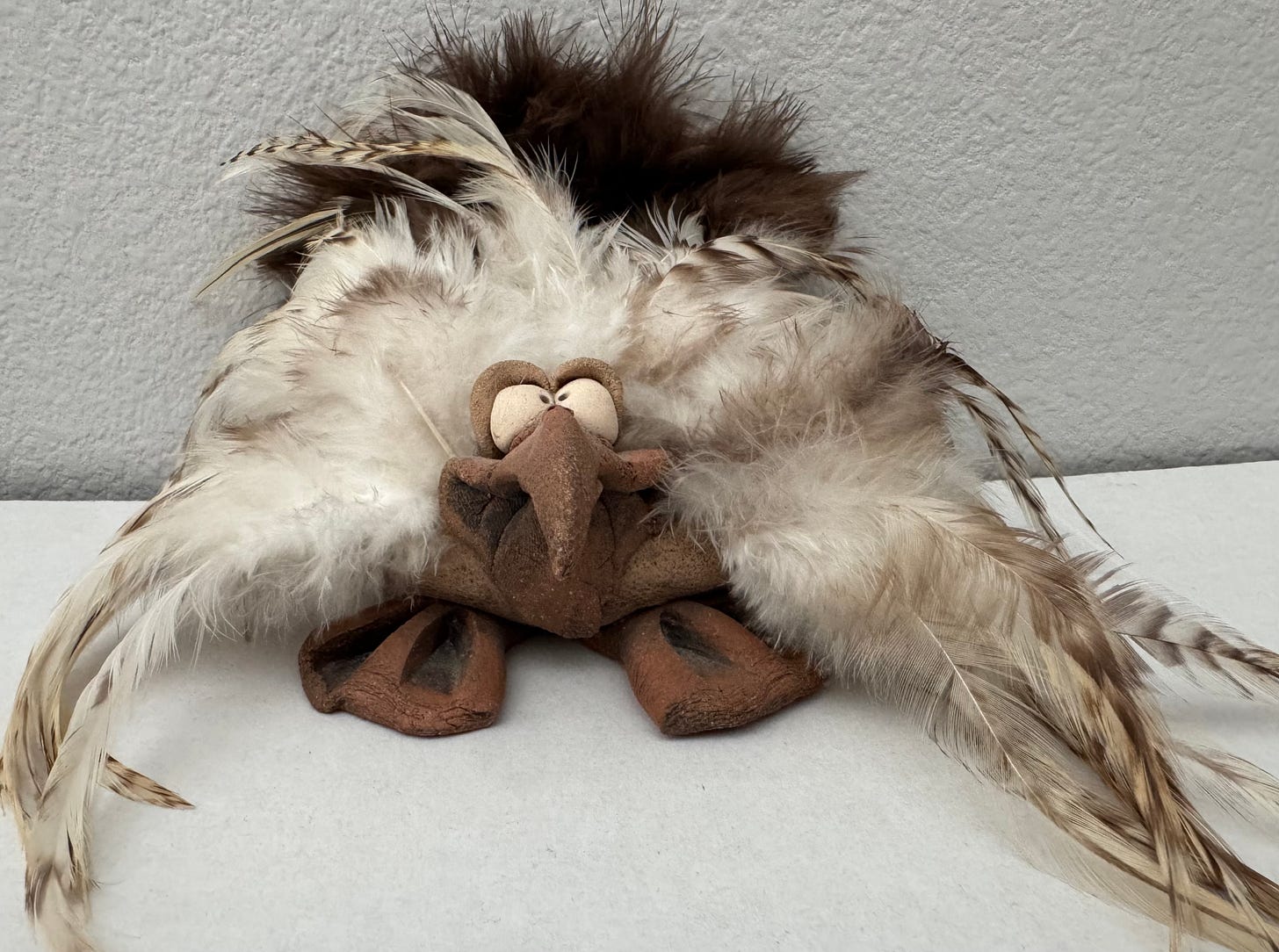 Brown clay bird with big white crossed eyes and duckish feet with wild white and brown feathers.