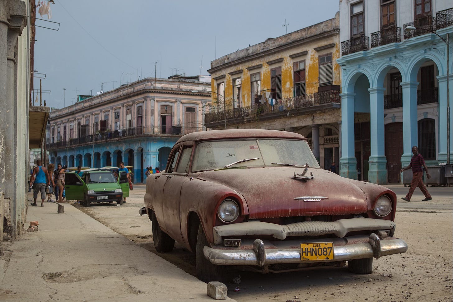 A weathered vintage American car sits parked on a dusty Havana street, its red paint faded and windshield clouded. Behind it, pastel buildings with peeling facades and hanging laundry line a sunlit block, while locals walk and gather nearby. Captures the layered textures of Cuba in 2014.