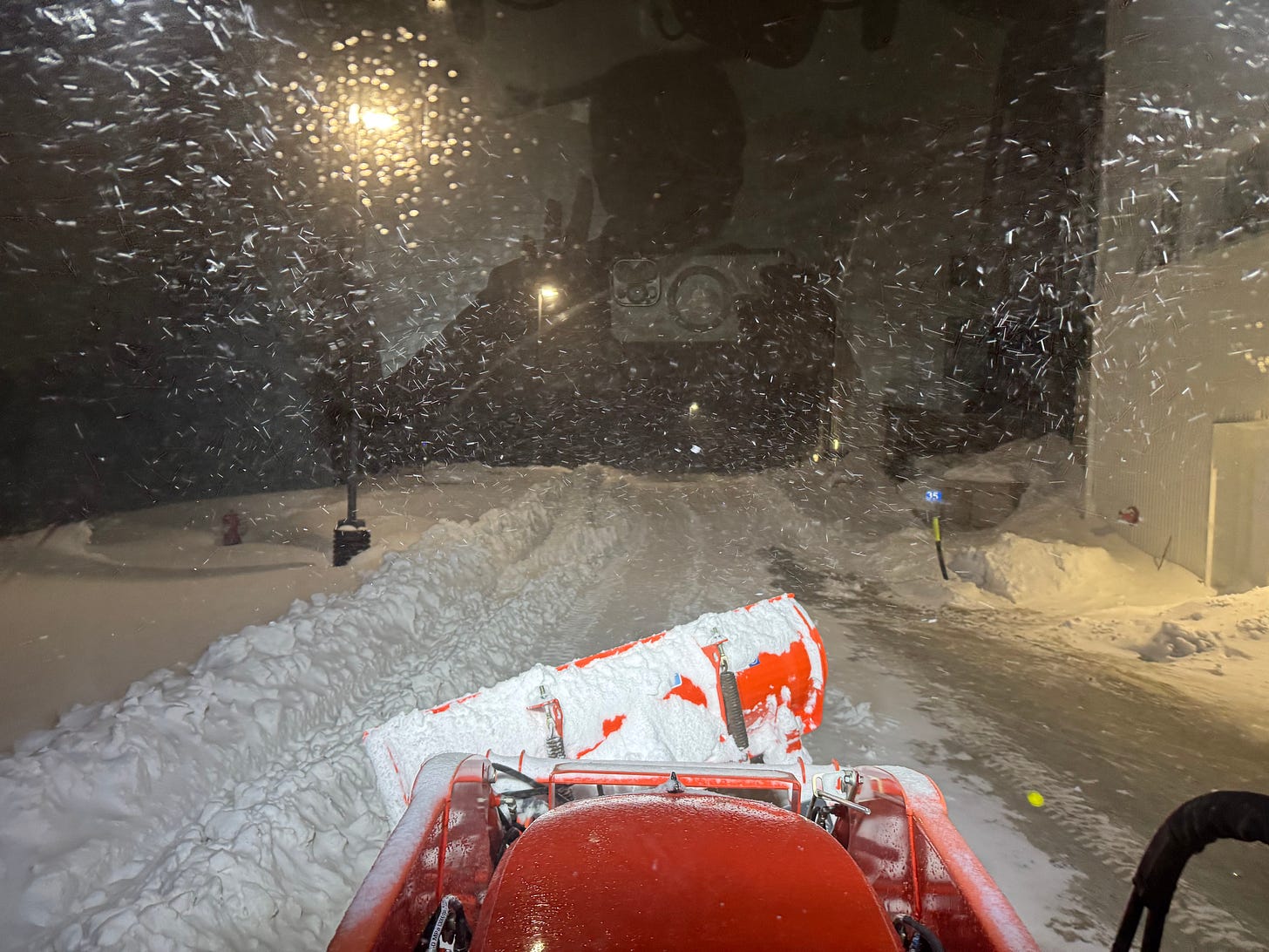 The view driving an orange tractor with a snow blade on the front, at night, it is snowing and there are three foot tall snowbanks. The view driving an orange tractor with a snow blade on the front, at night, it is snowing and there are three foot tall snowbanks.