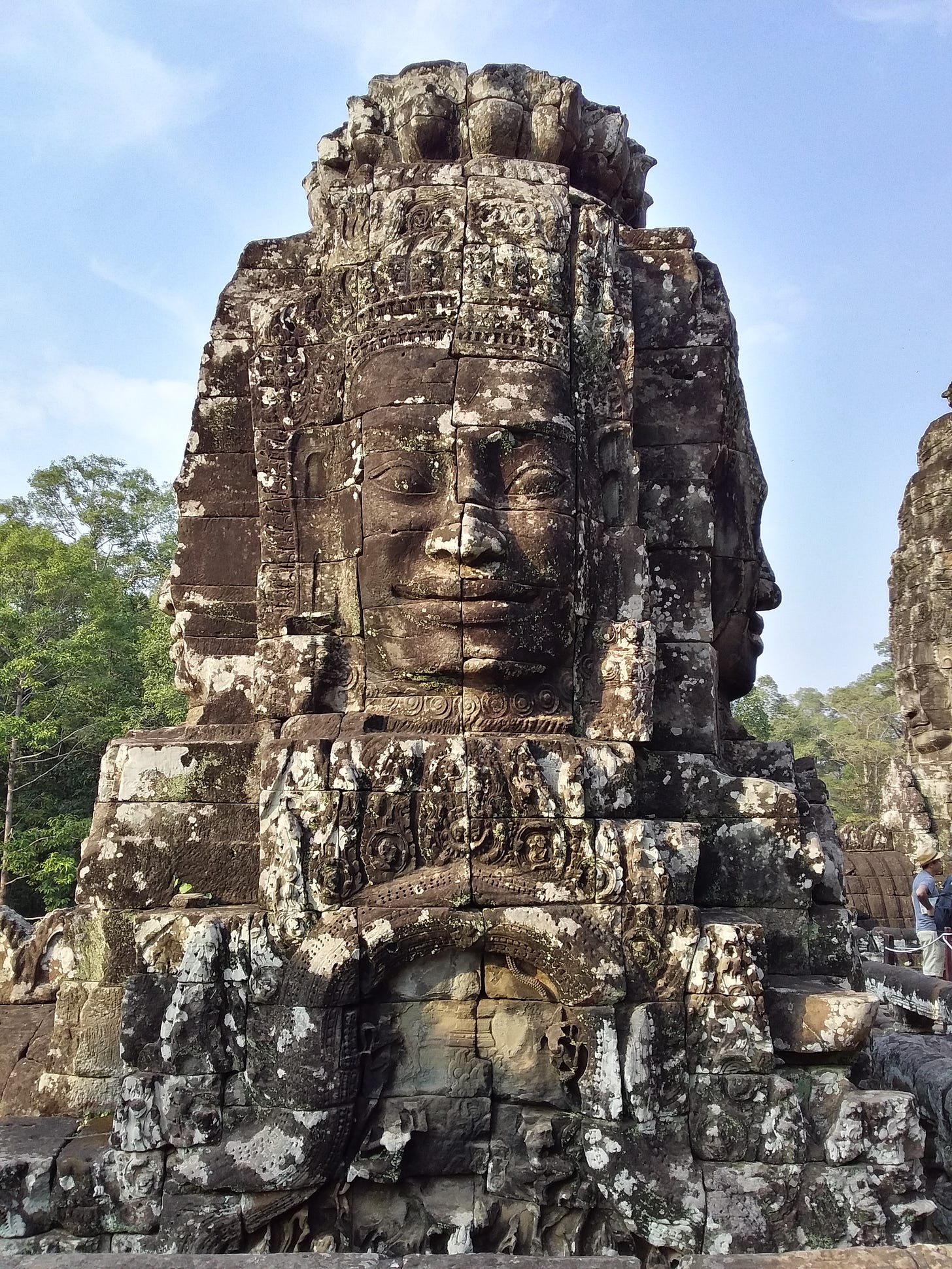 A close-up photograph of a massive, ancient stone tower at Bayon Temple in Cambodia. The tower features a giant, serene smiling face carved from large, weathered, lichen-covered stone blocks. The intricate ruins are set against a clear blue sky with green trees visible in the background.