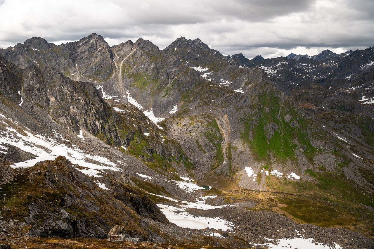 Looking down into a high mountain valley from the pass. Rows of jagged grey peaks recede into the distance under a heavy, overcast sky, their gullies streaked with snow. A green-and-scree valley floor curves below, holding a small blue-green tarn near a large snowfield. One hillside catches a band of sunlight through the cloud, glowing green against the surrounding grey. Looking down into a high mountain valley from the pass. Rows of jagged grey peaks recede into the distance under a heavy, overcast sky, their gullies streaked with snow. A green-and-scree valley floor curves below, holding a small blue-green tarn near a large snowfield. One hillside catches a band of sunlight through the cloud, glowing green against the surrounding grey.