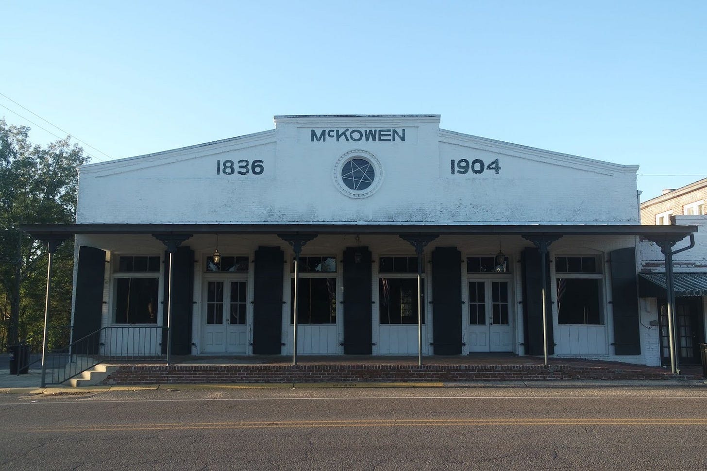 A historic white building with "McKOWEN" and dates "1836-1904" painted on its front facade, featuring a covered porch with dark columns and multiple doors along a street.