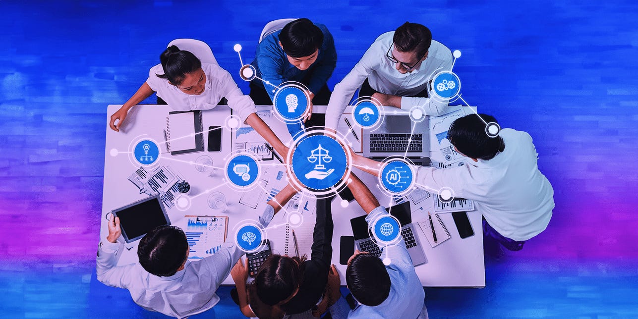 A graphic from above shows several people in corporate attire gathered around a table with laptops, notebooks, and stationery on it. They point to the center, where a large blue circular graphic with a symbol connecting to other graphic symbols scattered around it. A graphic from above shows several people in corporate attire gathered around a table with laptops, notebooks, and stationery on it. They point to the center, where a large blue circular graphic with a symbol connecting to other graphic symbols scattered around it.