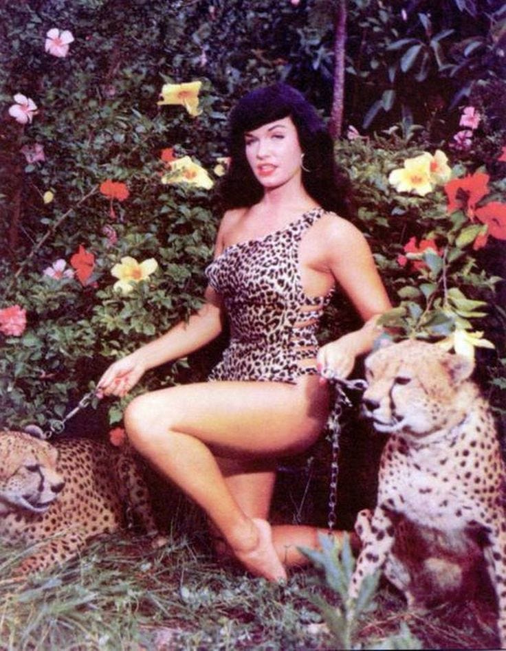This may contain: a woman sitting on the ground with two cheetah in front of flowers and bushes