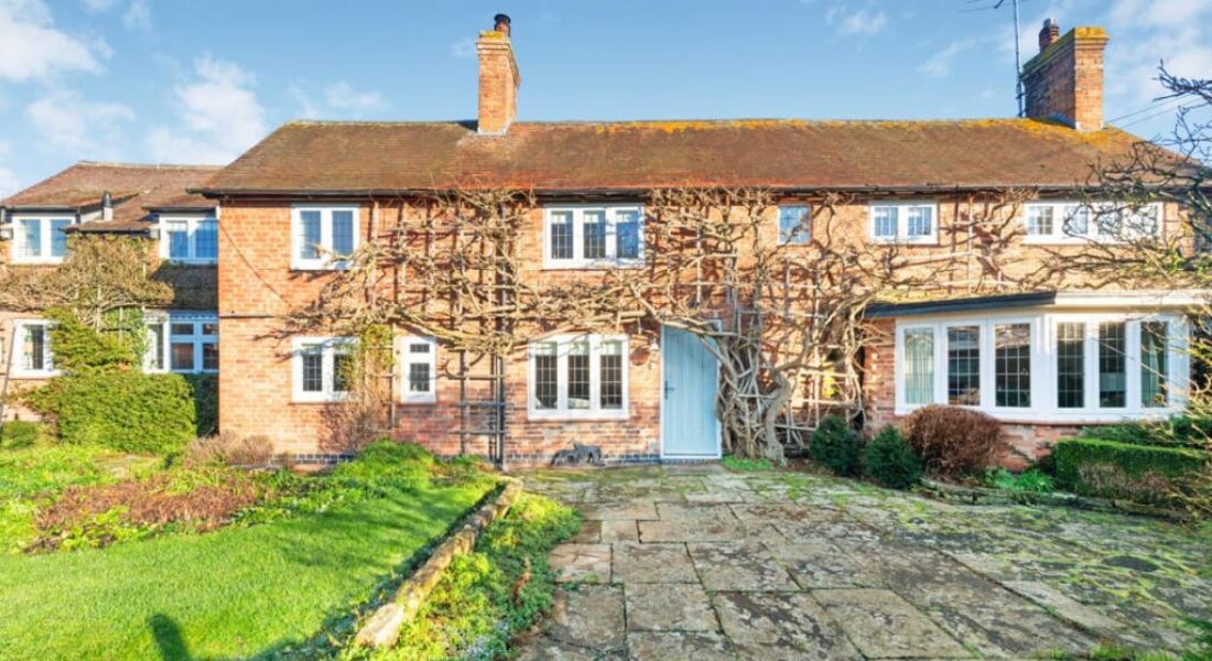 A large brick cottage with white windows, a blue door, and a lawn and a concrete driveway in front of it