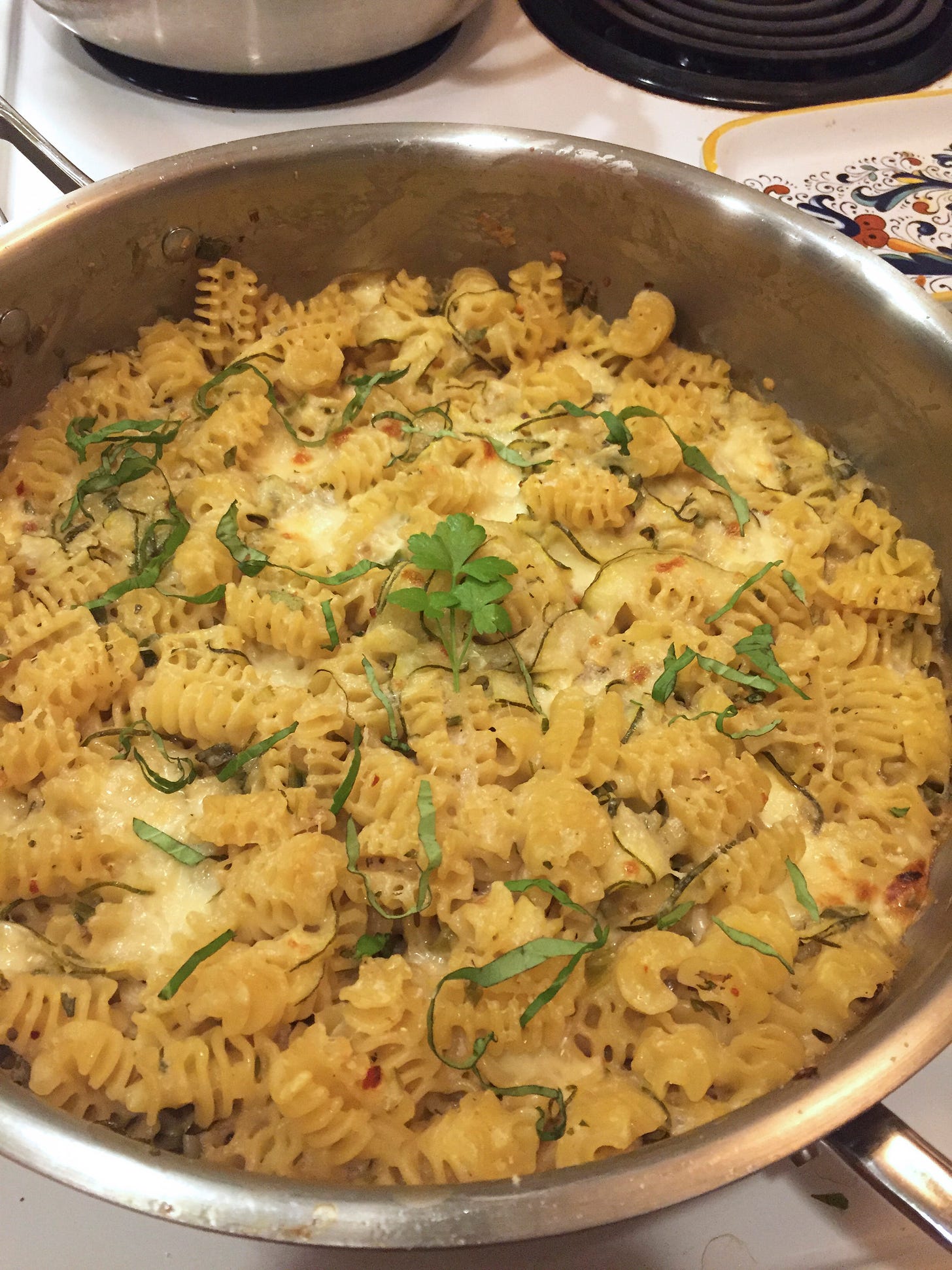 On the stove is a steel pan filled with a cheesy radiatore dish, with slices of zucchini throughout. On top is a sprig of Italian parsley and pieces of fresh basil.