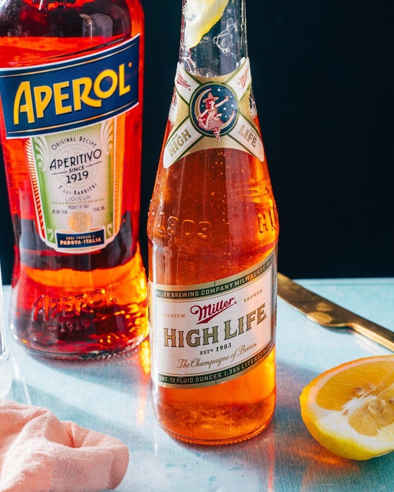 A bottle of Miller High Life beer next to a bottle of Aperol and a wedge of lime