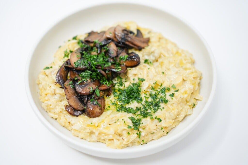 A bowl of creamy orzo pasta mixed with artichoke hearts, topped with golden-brown caramelized mushrooms, and garnished with fresh parsley.