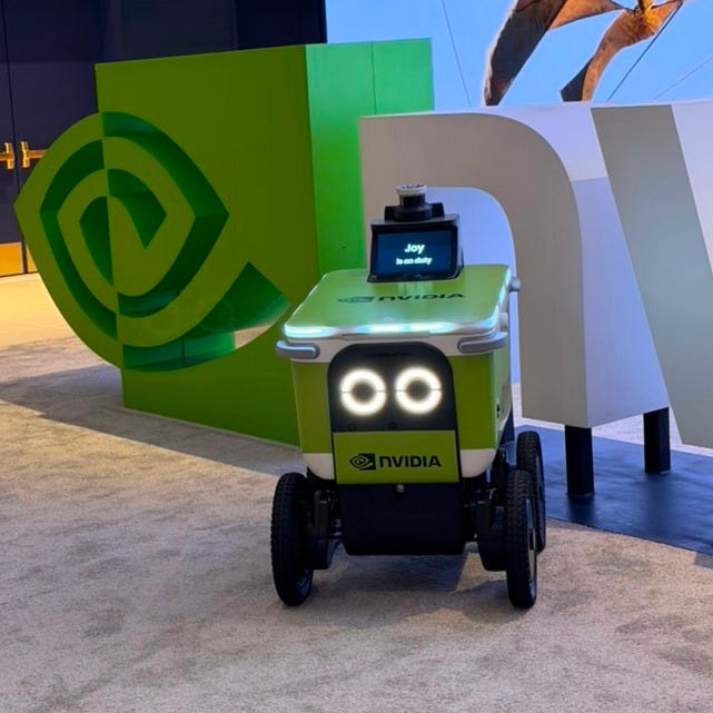 A small NVIDIA delivery robot with glowing circular headlights stands in front of a large NVIDIA logo display at CES. A screen on top reads “Joy is on duty.” A small NVIDIA delivery robot with glowing circular headlights stands in front of a large NVIDIA logo display at CES. A screen on top reads “Joy is on duty.”