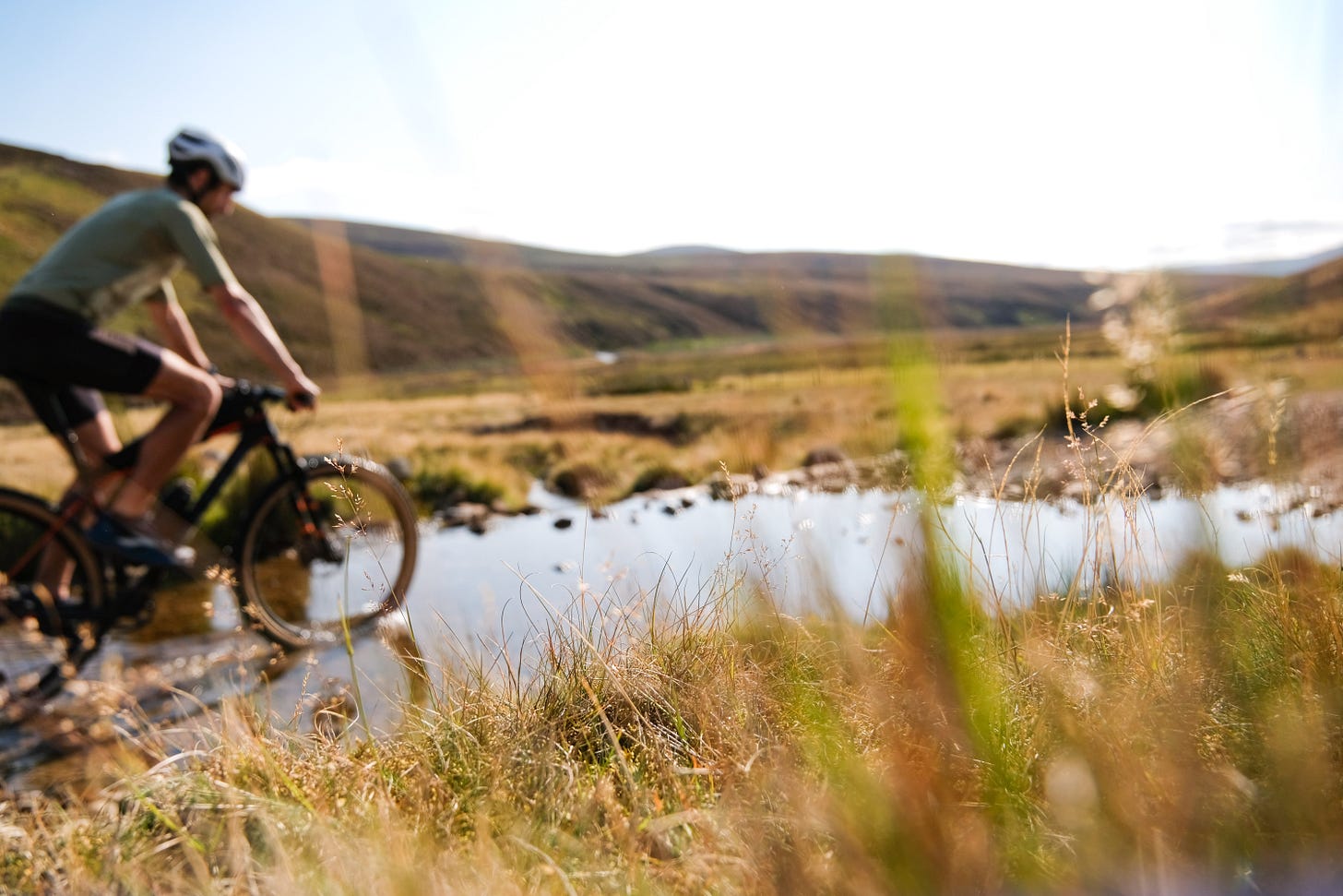 A cyclist rides through a shallow river, blurred in the background. In sharp focus are the blades of grass in the foreground, highlighting the contrast between sweeping Highland landscapes and the intimate details of nature.