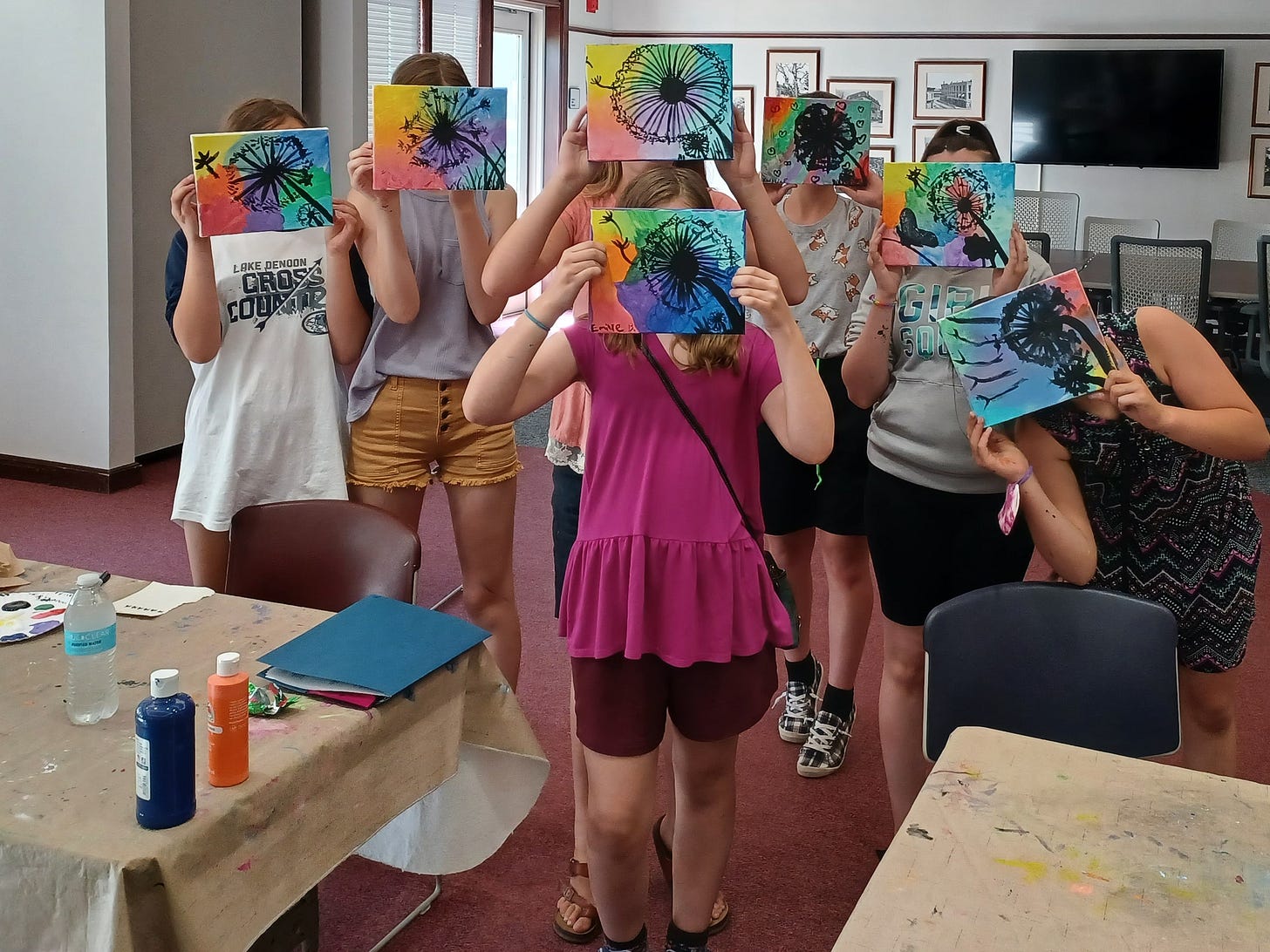 A group of young people hold colorful artworks over their faces in a library makerspace. A group of young people hold colorful artworks over their faces in a library makerspace.