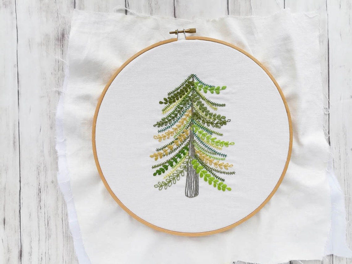 May include: A green, yellow, and brown embroidered tree design on white fabric in a wooden embroidery hoop.