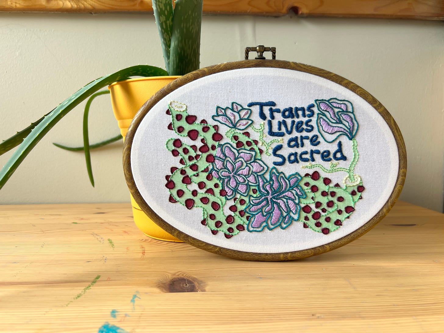 Completed mixed-media embroidery piece using the design created by Ris Wong of winding thorny vines and blossoming many-petalled flowers. The piece is leaning up against a yellow plant pot with an Aloe plant growing in it. Completed mixed-media embroidery piece using the design created by Ris Wong of winding thorny vines and blossoming many-petalled flowers. The piece is leaning up against a yellow plant pot with an Aloe plant growing in it.