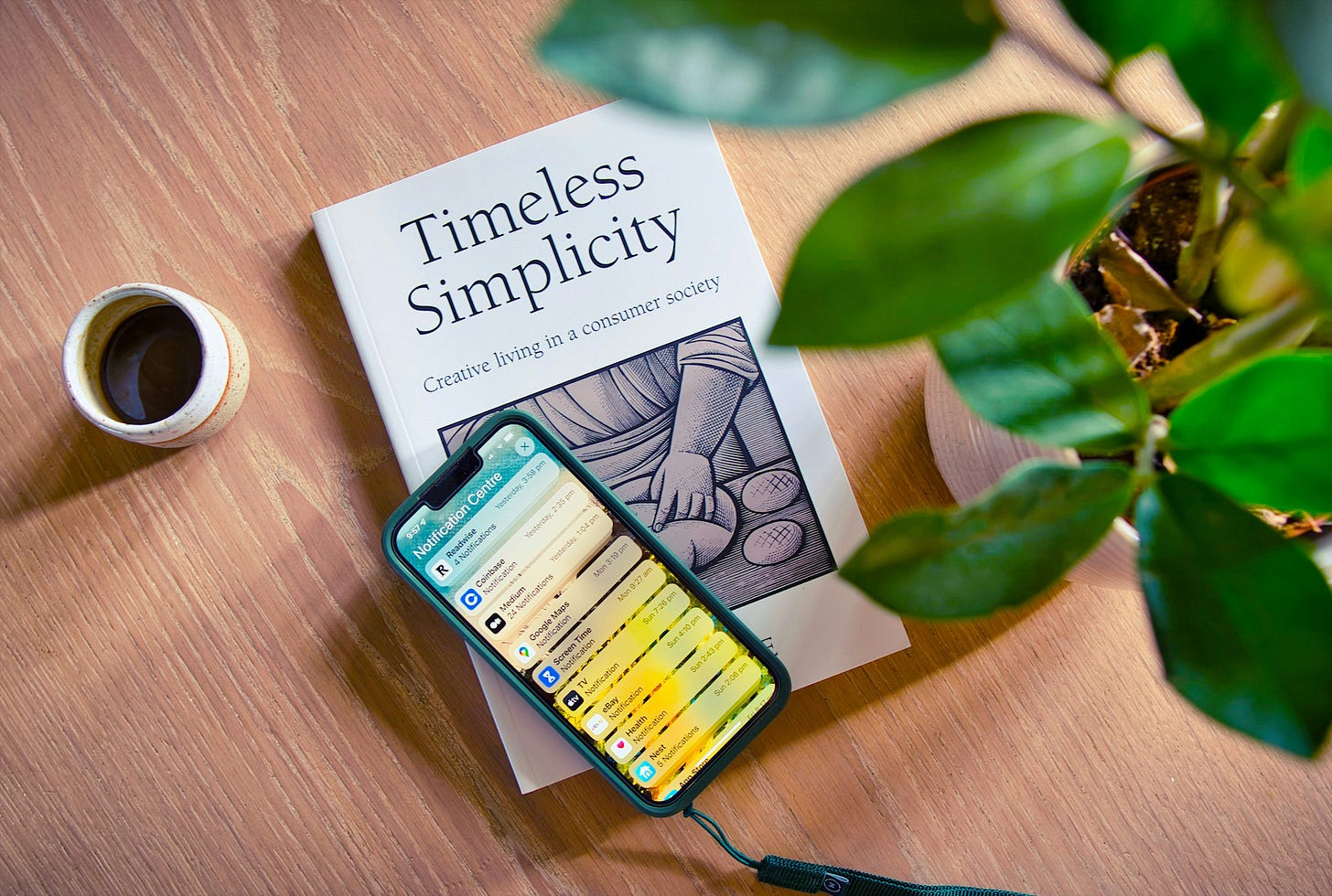 Book titled "Timeless Simplicity" on a wooden table with a smartphone displaying notifications and a cup of coffee; lush green plant nearby. Cozy, peaceful setting.