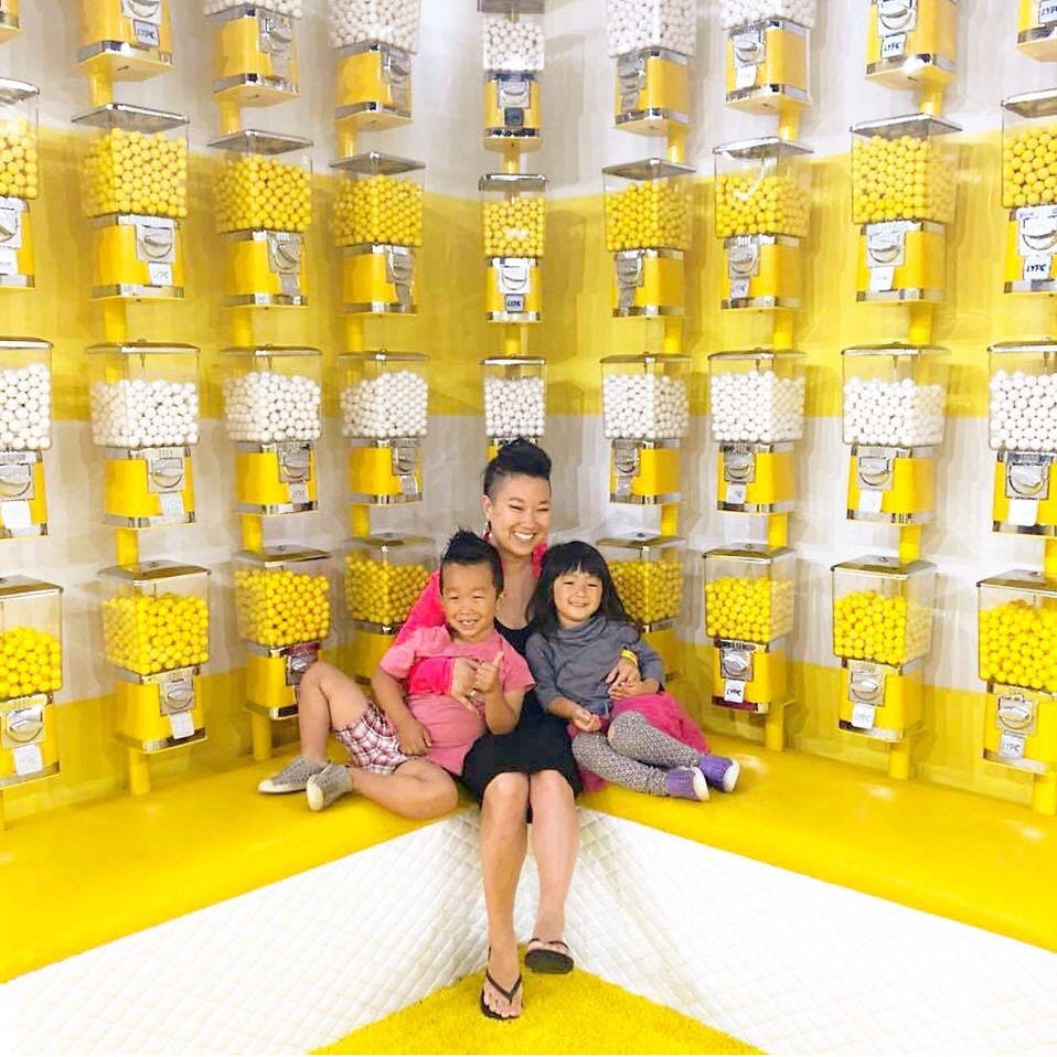 Cancer survivor Sarah smiles with two kids in a bright room filled with yellow and white gumball machines Cancer survivor Sarah smiles with two kids in a bright room filled with yellow and white gumball machines
