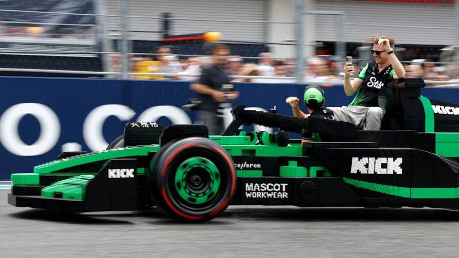 A life-sized LEGO Formula 1 car in Green and Black is being driven on a racetrack with 2 people inside, and a crowd in the background.