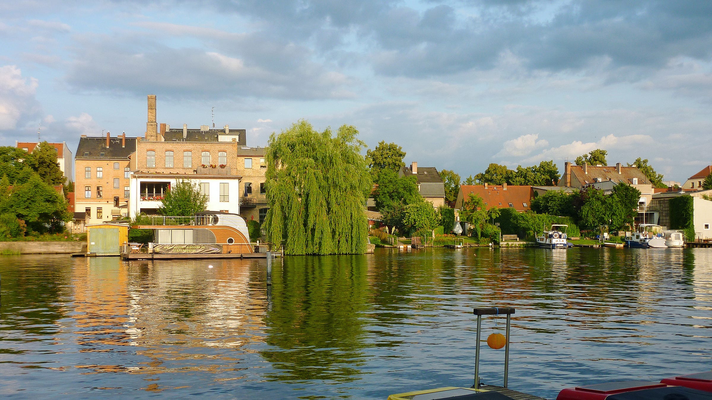 A row of brick buildings along a riverfront, photographed at sunset in the spring. A small houseboat is in the left foreground. A row of brick buildings along a riverfront, photographed at sunset in the spring. A small houseboat is in the left foreground.