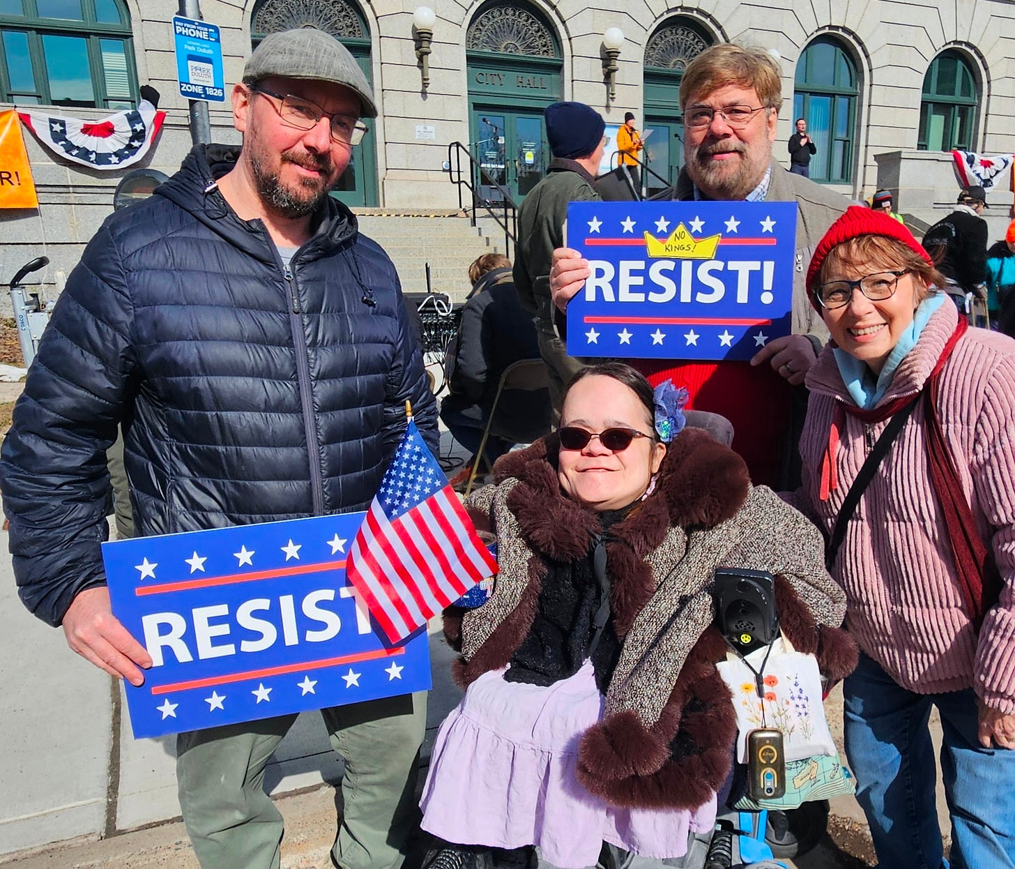 A picture of Gaelynn and Paul and her parents standing in front of City Hall in Duluth during the last No Kings Rally. Paul is holding a resist sign and Gaelynn's Dad is holding a Resist Sign and Gaelynn's Mom is wearing a red knit cap.