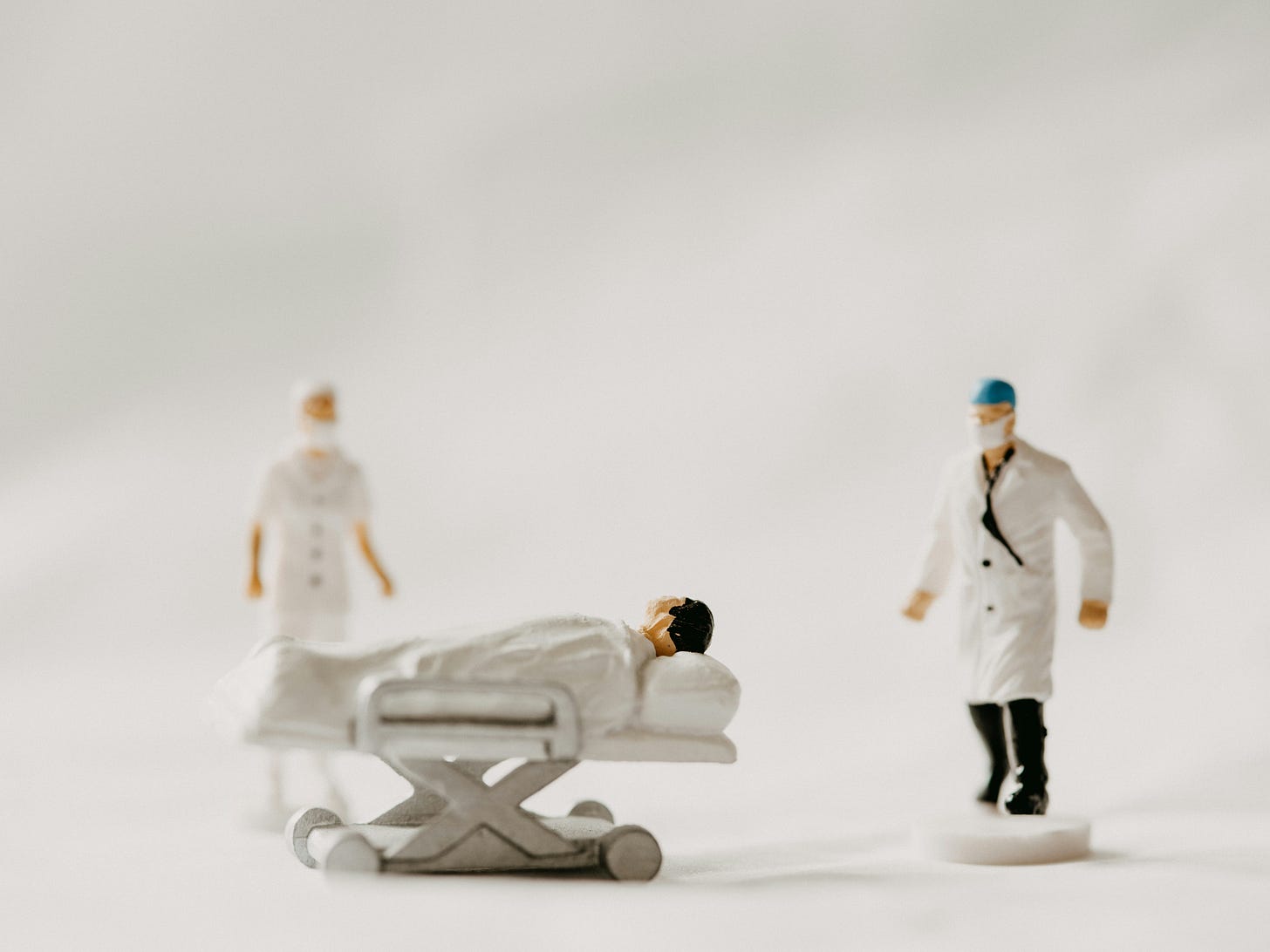 Small medical figurines. On the left is a nurse, in the middle is a patient on a gurney, and there’s a doctor on the right wearing a mask and skull cap.