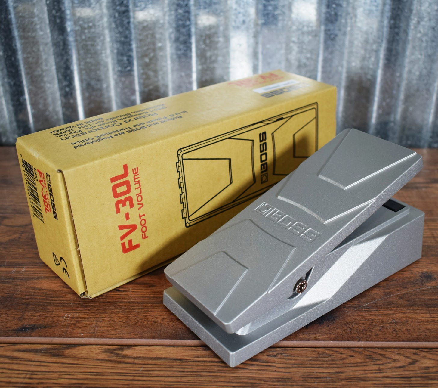 A silver BOSS FV-30L foot volume pedal resting on a wooden table beside its yellow box, photographed against a corrugated metal backdrop.