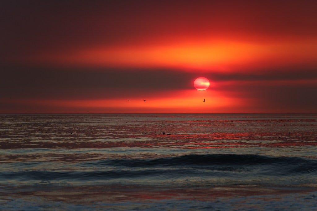 A view of a fiery red sun setting over the ocean as smoke fills the air. A view of a fiery red sun setting over the ocean as smoke fills the air.